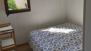 A small, sparsely furnished room with a single bed, a wooden shelf unit, and a window providing a view of the outside.
