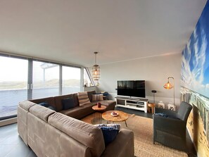 A modern living room with a sectional sofa, a flat-screen TV, and a large window with blinds. - Callantsoger Staete A402 (Callantsoog)