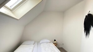Une petite chambre avec un lit simple, une lucarne, une table de chevet avec une lampe et un objet noir accroché au mur.