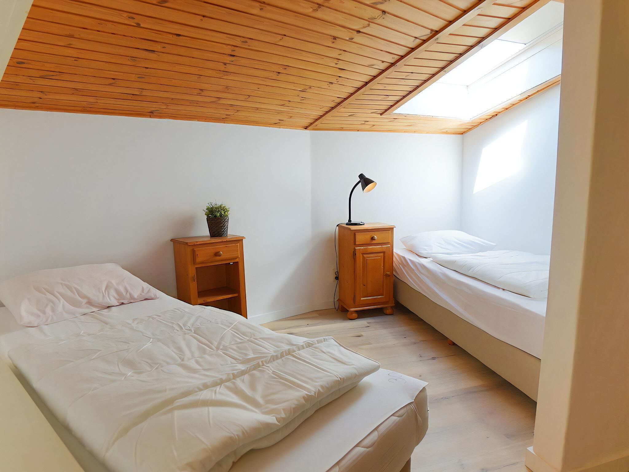 Une petite chambre bien éclairée, avec deux lits, une table de chevet en bois surmontée d’une lampe et une lucarne.