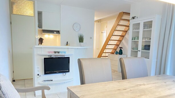 A modern kitchen with a built-in TV, white cabinets, and a staircase leading to a dining area with a wooden table and chairs. - 2 bedroom accommodation in Callantsoog (Callantsoog)