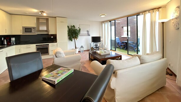 Ein modernes Wohnzimmer mit einer Couch, einem Couchtisch und einem Esstisch. Eine Küchenzeile mit Schränken und einer Mikrowelle ist ebenfalls vorhanden. Ein großes Fenster bietet einen Blick nach außen.