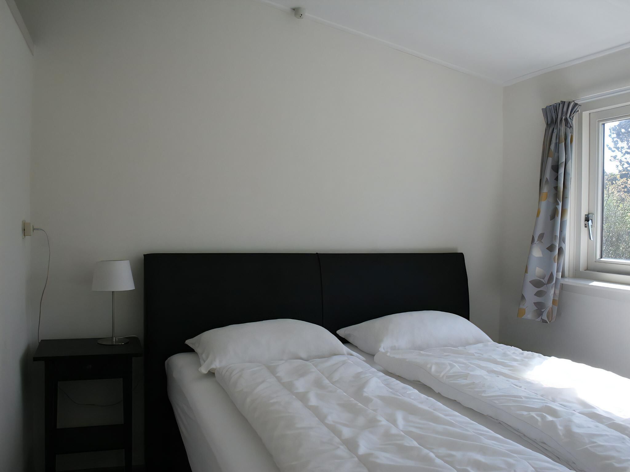Ein schlichter, sauberer Raum mit einem Bett, einem Nachttisch mit Lampe und einem mit Vorhängen versehenen Fenster.
