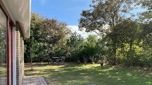 A patio with a brick floor, a glass door, and a view of a grassy yard with trees and a wooden table. - 2 bedroom accommodation in Callantsoog (Callantsoog)