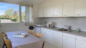 A modern kitchen with white cabinets, a dining table with chairs, a coffee maker, and a window with curtains. - 2 bedroom accommodation in Callantsoog (Callantsoog)