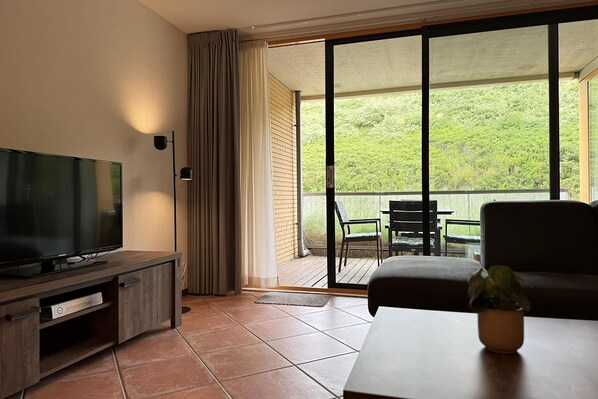 Une pièce avec un téléviseur posé sur un meuble en bois, un canapé, une table avec une plante et un balcon donnant sur un paysage verdoyant.