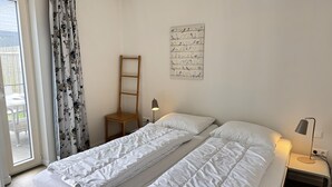 Een slaapkamer met een bed, nachtkastje, lamp, raam met gordijnen en een aan de muur hangend vogelillustratie.