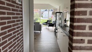 Een smalle gang die uitkomt op een keuken met een bakstenen muur, een eethoek met stoelen en een uitzicht op een huis op de achtergrond.