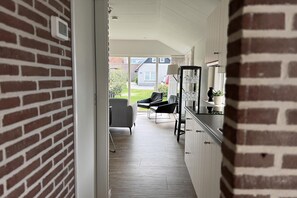 A narrow hallway leading to a kitchen with a brick wall, a dining area with chairs, and a view of a house in the background. - 2 bedroom accommodation in Callantsoog (Callantsoog)