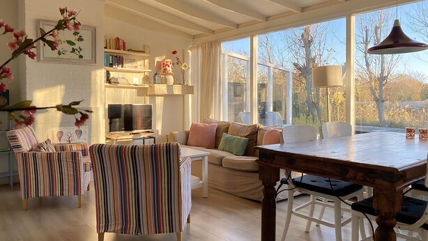 A cozy living room with a striped armchair, a sofa, a dining table, and a TV. There are bookshelves and a window with a view of trees. - 3 bedroom accommodation in Callantsoog (Callantsoog)