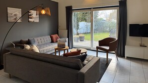 Ett modernt vardagsrum med en grå soffa, ett soffbord och en tv. Det finns en stor skjutdörr i glas som leder ut till en trädgård.