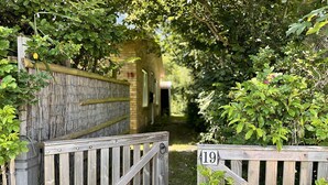 A wooden gate with a number sign, leading to a brick building surrounded by lush greenery.
