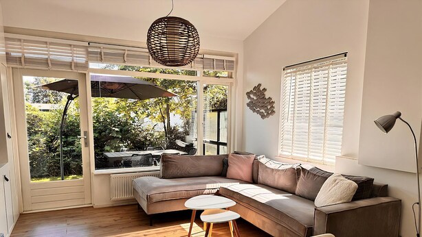 A modern living room with a sectional sofa, a round coffee table, and a chair. There is a large window with blinds, a hanging light fixture, and a radiator.