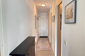 A narrow hallway with a wooden floor, a black bench on the left, and a door at the end.