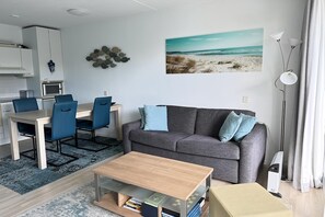 A modern living room with a grey sofa, blue chairs, a wooden coffee table, and a beach-themed painting on the wall.