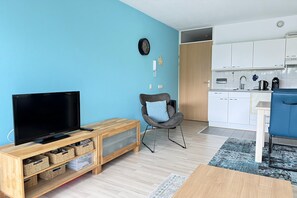 A modern living room with a television, a chair, and a kitchen area with cabinets and a sink. - 1 bedroom accommodation in Callantsoog (Callantsoog)