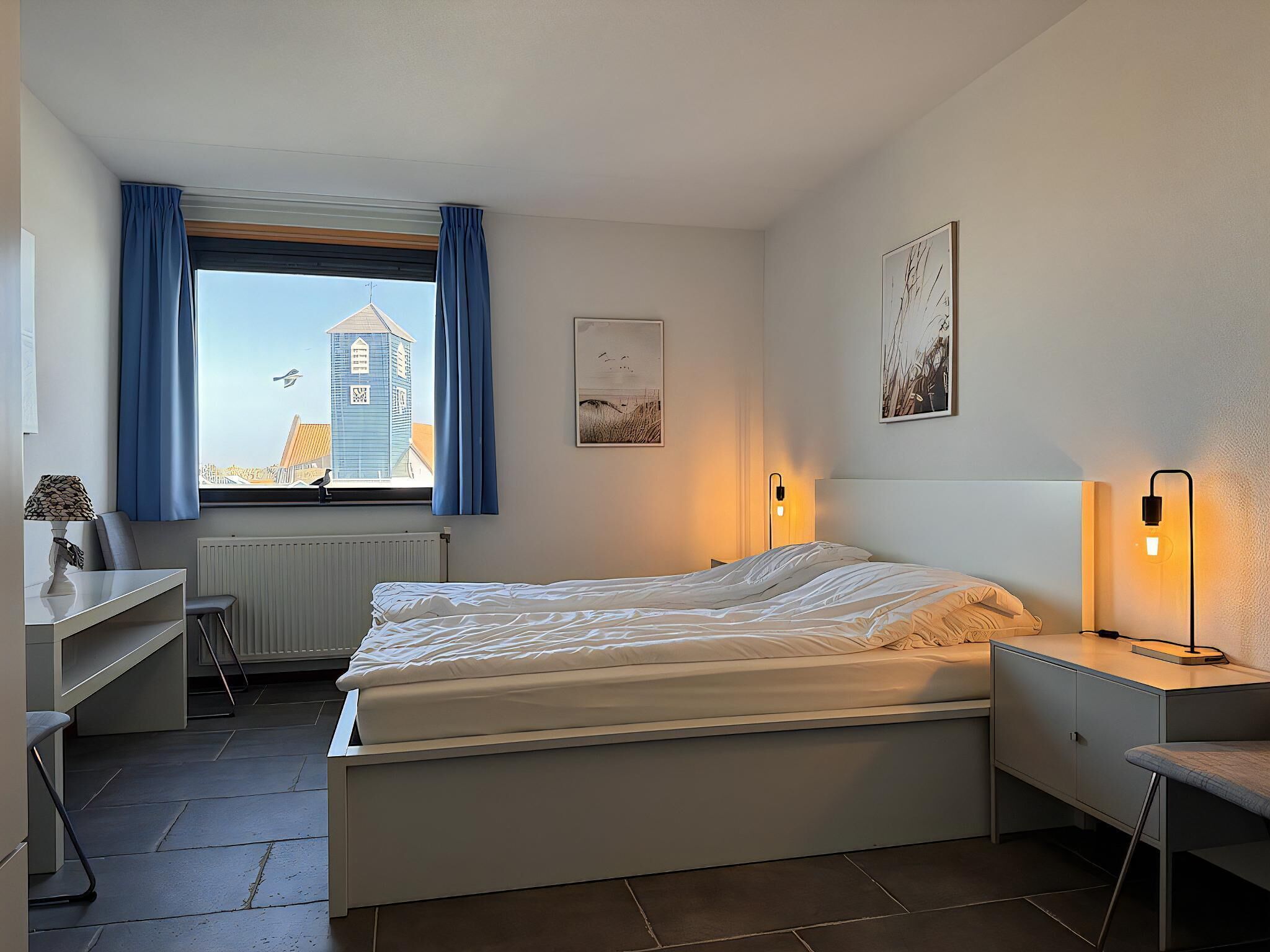 Een slaapkamer met een bed, een bureau, een stoel, een radiator, een raam met gordijnen en kunstwerken aan de wanden.