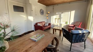A cozy living room with a wooden ceiling, a red sofa, a black chair, and a dining table with a magazine.
