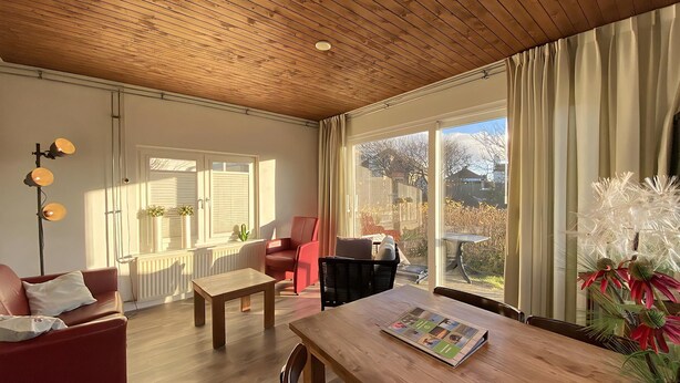 A spacious living room with a wooden ceiling, a dining table with a magazine, a red armchair, and a large window with curtains.