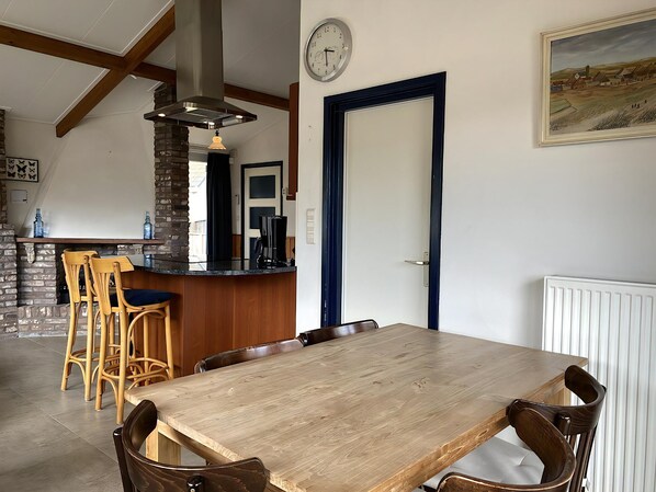 A dining area with a wooden table and chairs, a kitchen with a brick wall and a hanging light fixture, and a clock on the wall.