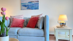 A living room with a grey sofa, red and striped pillows, a painting of a coastal scene, a lamp, and a potted plant. - Callantsoger Staete A306 (Callantsoog)
