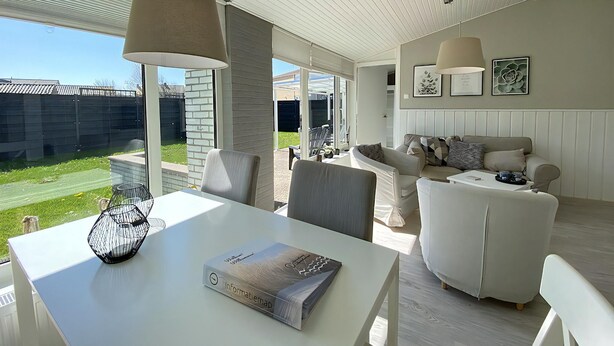 A modern living room with a white sofa, a dining table with a book, and a large window overlooking a garden. - 3 bedroom accommodation in Callantsoog (Callantsoog)