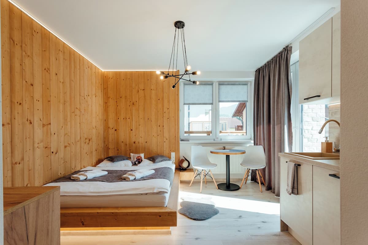 A modern bedroom with a wooden wall, a bed with pillows and a blanket, a bedside table, a small round table with chairs, a window with curtains, and a kitchen area with a sink and cabinets.