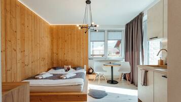 A modern bedroom with a wooden wall, a bed with pillows and a blanket, a bedside table, a small round table with chairs, a window with curtains, and a kitchen area with a sink and cabinets.