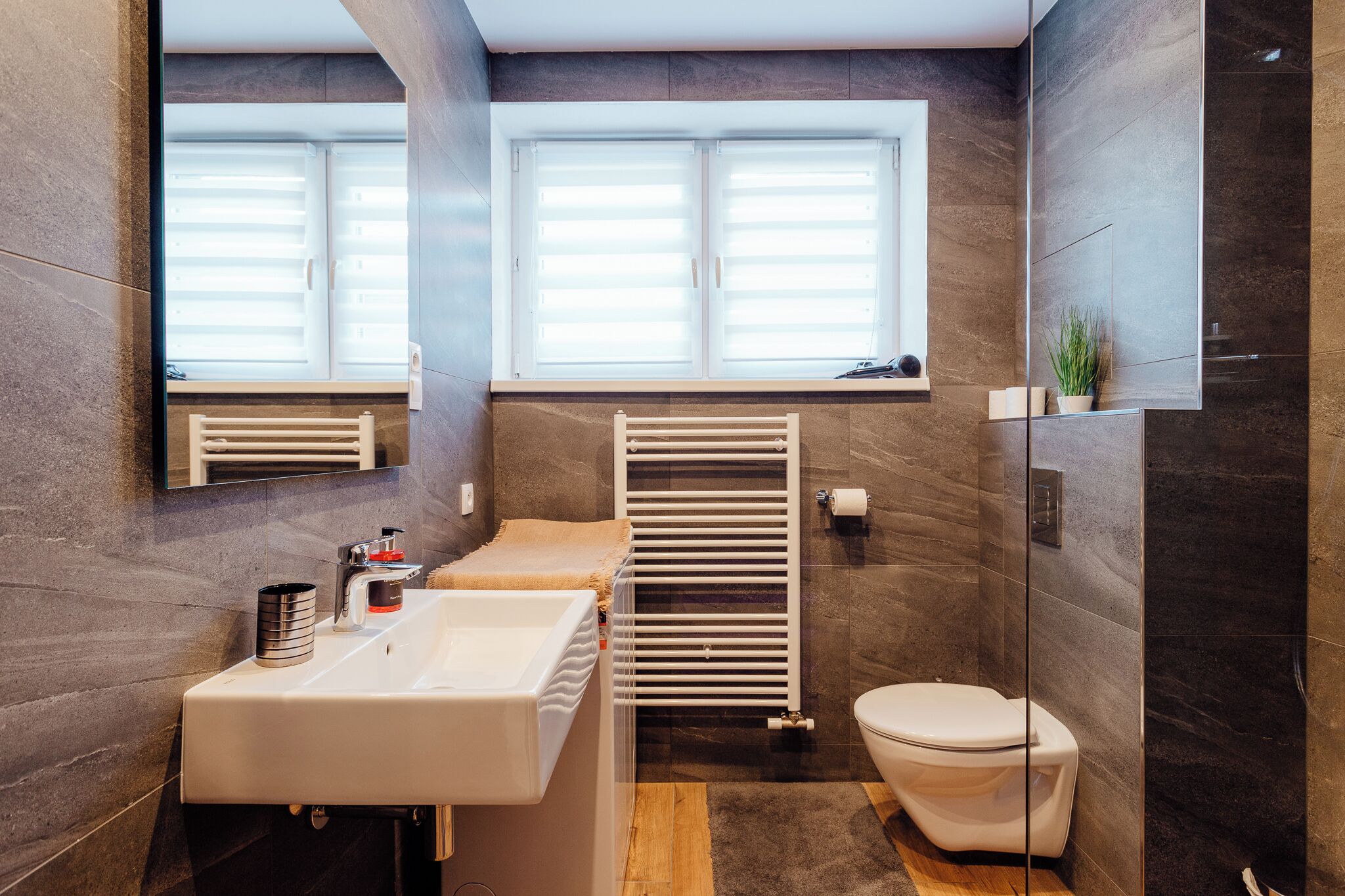 A modern bathroom with a white sink, a toilet, a towel rack, and a window with blinds.