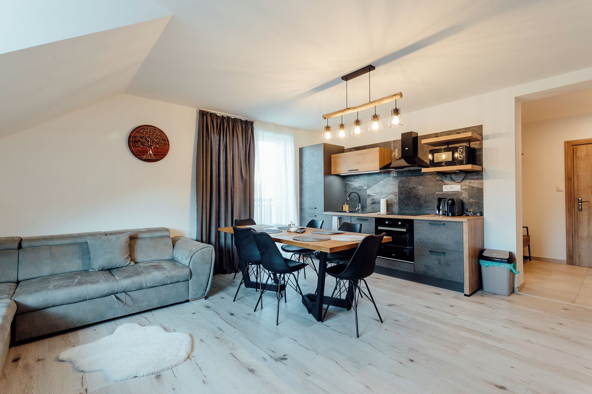 Sala de estar moderna com sofá cinza, mesa de jantar e cadeiras. Cozinha com micro-ondas, fogão e armários visível.