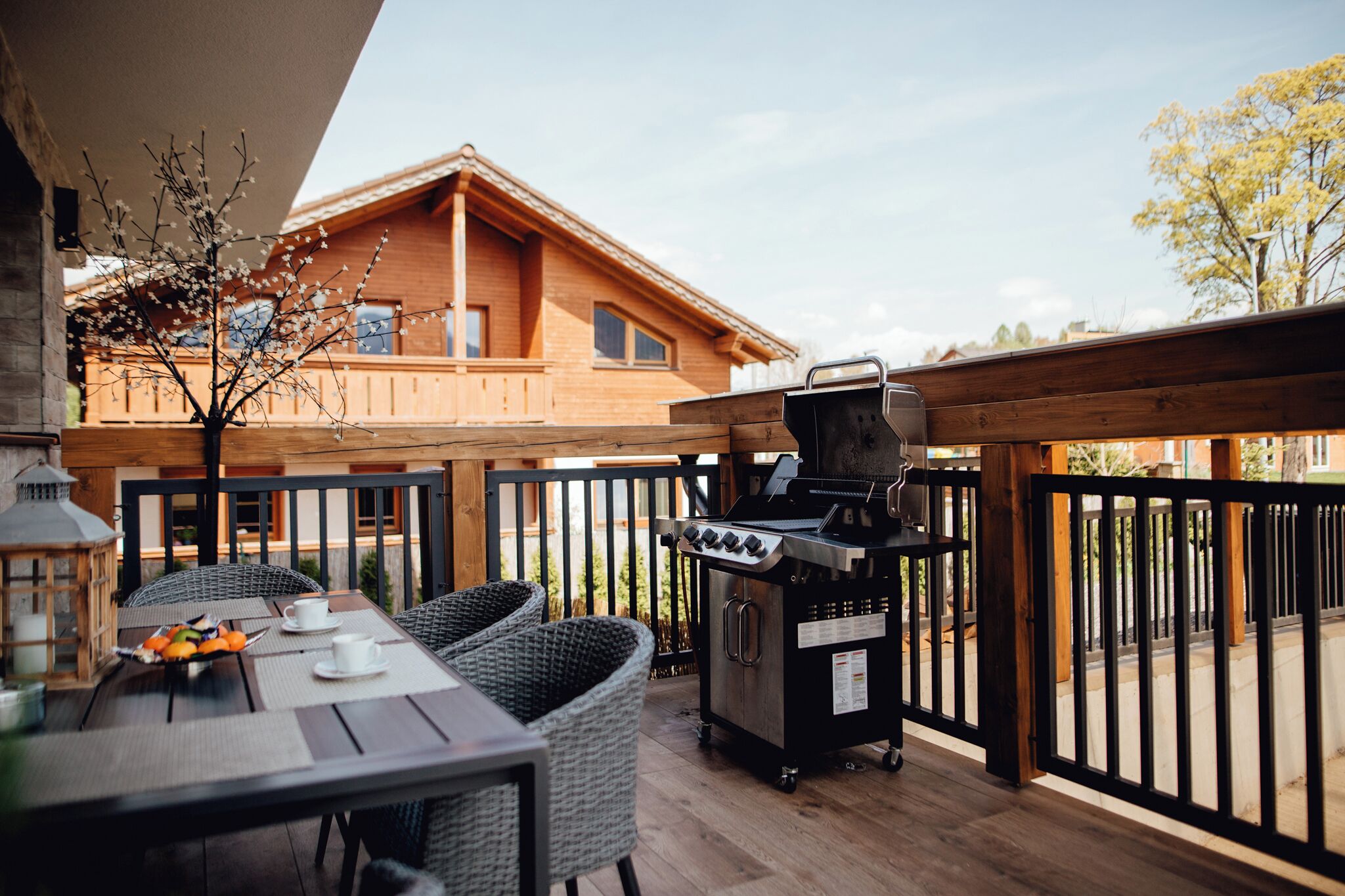Ein Essbereich im Freien mit Korbstühlen und einem Grill, einer Holzterrasse und Blick auf ein Holzgebäude im Hintergrund.