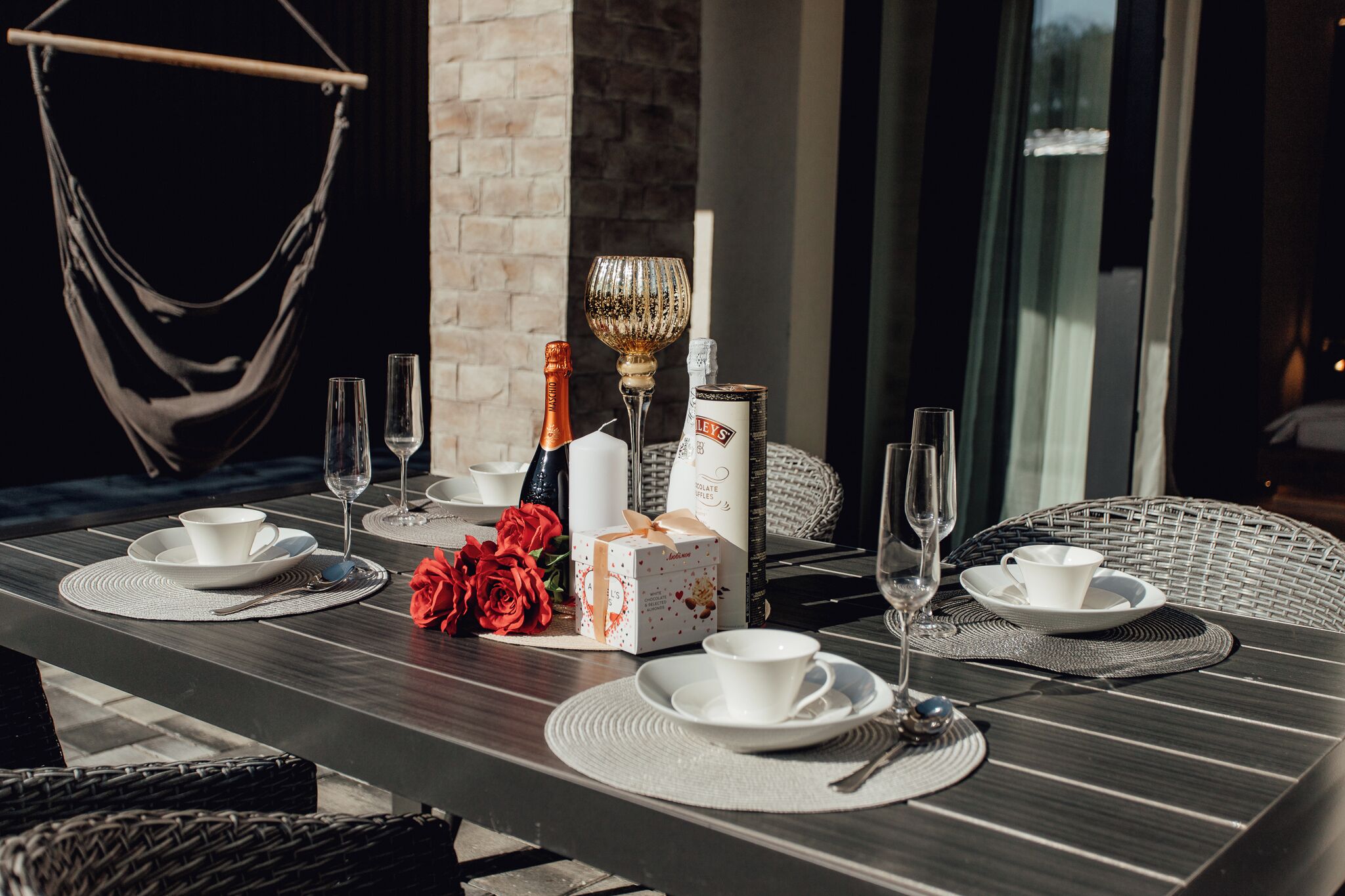 A dining table set with white plates, glasses, and a bottle of champagne. There are red roses and a gift box on the table.