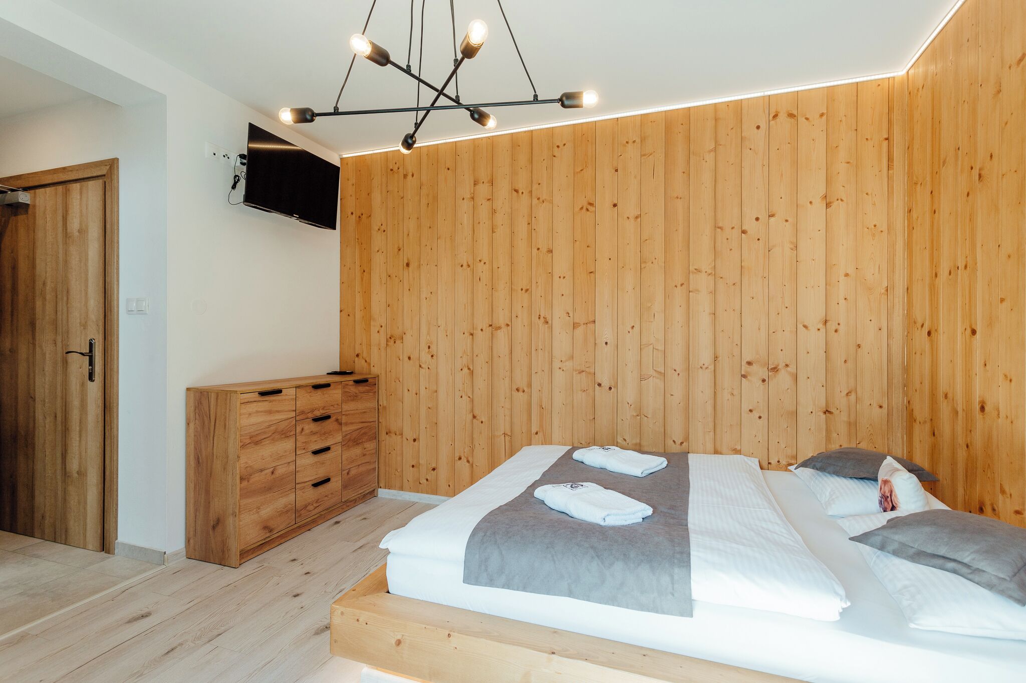 A modern bedroom with a wooden wall panel, a bed with a grey blanket, a wooden dresser, and a ceiling light fixture.