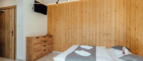 A modern bedroom with a wooden wall panel, a bed with a grey blanket, a wooden dresser, and a ceiling light fixture.