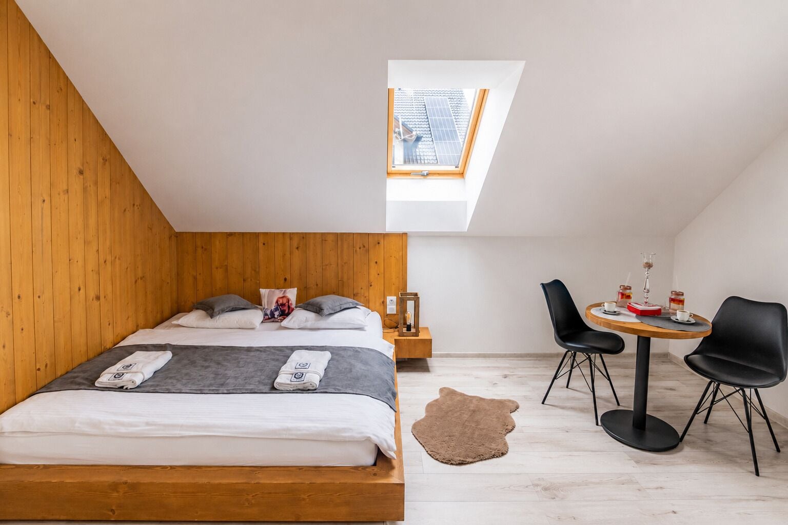 Un dormitorio con techo inclinado, tragaluces, cama con almohadas, mesita de noche con lámpara, una pequeña mesa redonda con sillas y una alfombra.