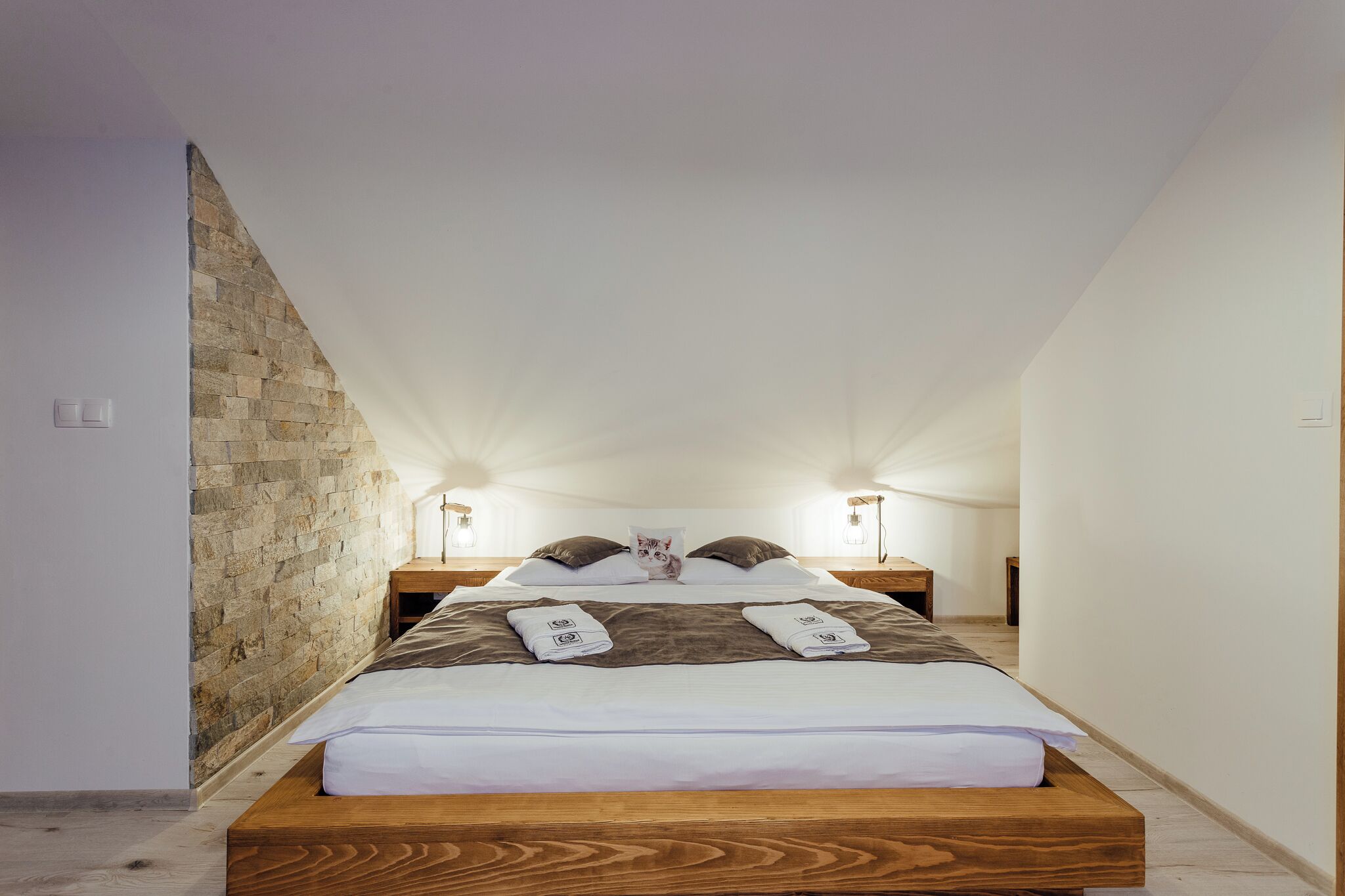 Een smalle, schuine kamer met een centraal geplaatst bed, een stenen muur aan de zijkant en twee nachtkastjes met lampen.