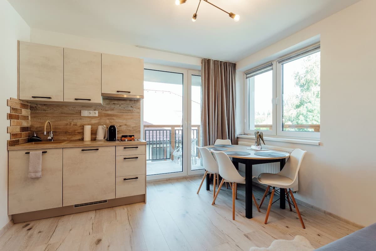 A modern kitchen with wooden cabinets, a dining area with a round table and chairs, and a view of the outdoors through large windows.