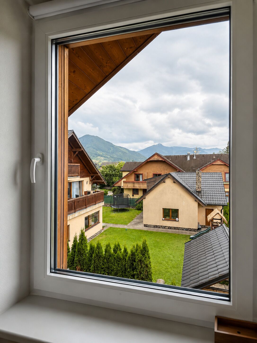 A window view of a residential area with houses, a grassy yard, and mountains in the background.