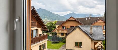 A window view of a residential area with houses, a grassy yard, and mountains in the background.