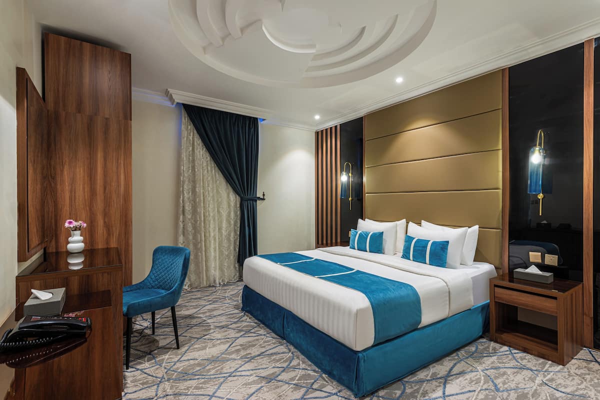 A hotel room with a large bed, a desk with a chair, a vanity with a mirror, and a nightstand with a lamp.