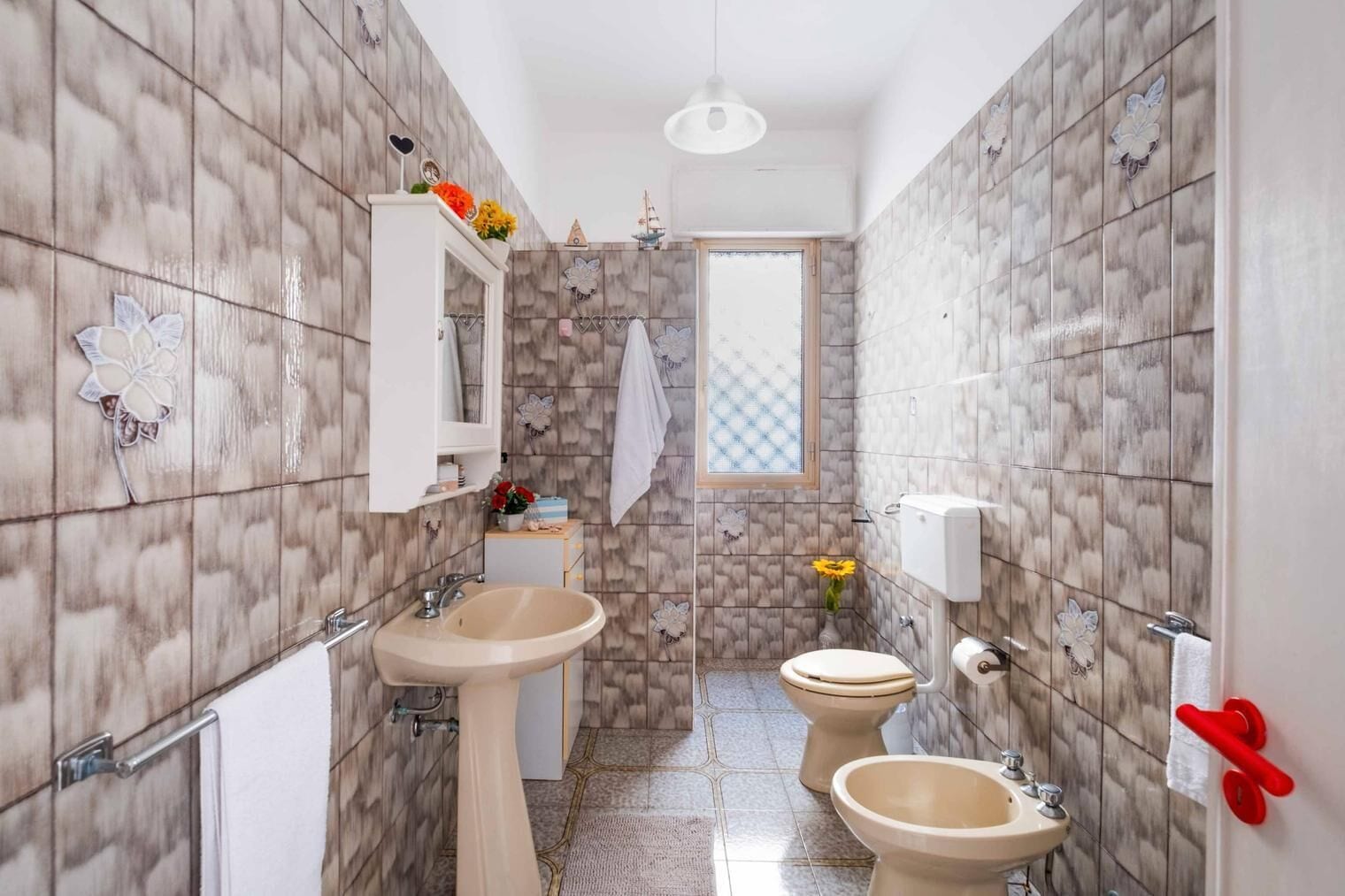 A bathroom with two sinks, a toilet, and a bidet. It has a window with a view, a mirror, and decorative tiles.