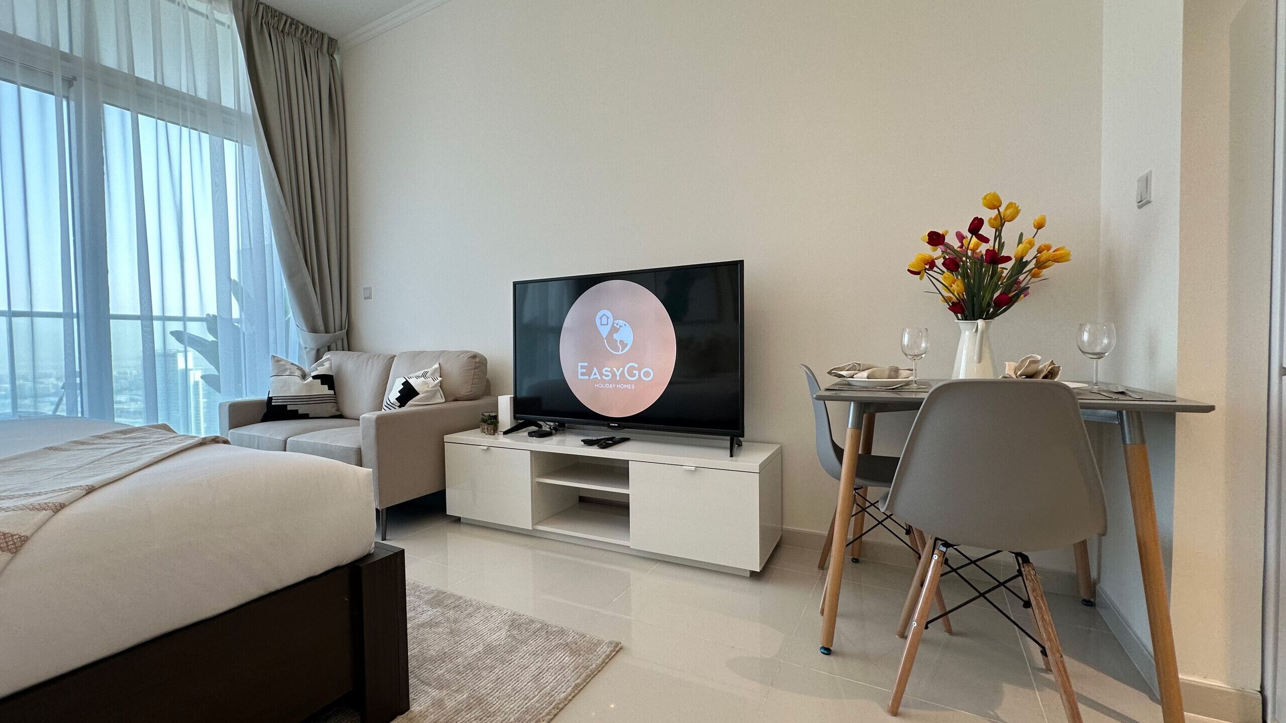 A modern hotel room with a bed, a TV on a white stand, a dining table with chairs, and a vase of flowers.