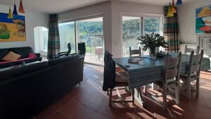 A spacious living area with a dark-colored sofa, a dining table with chairs, and a large window offering a view of greenery outside. - Casa Vale vinagre - Villa large pool for max. 8 persons (São Bartolomeu de Messines)