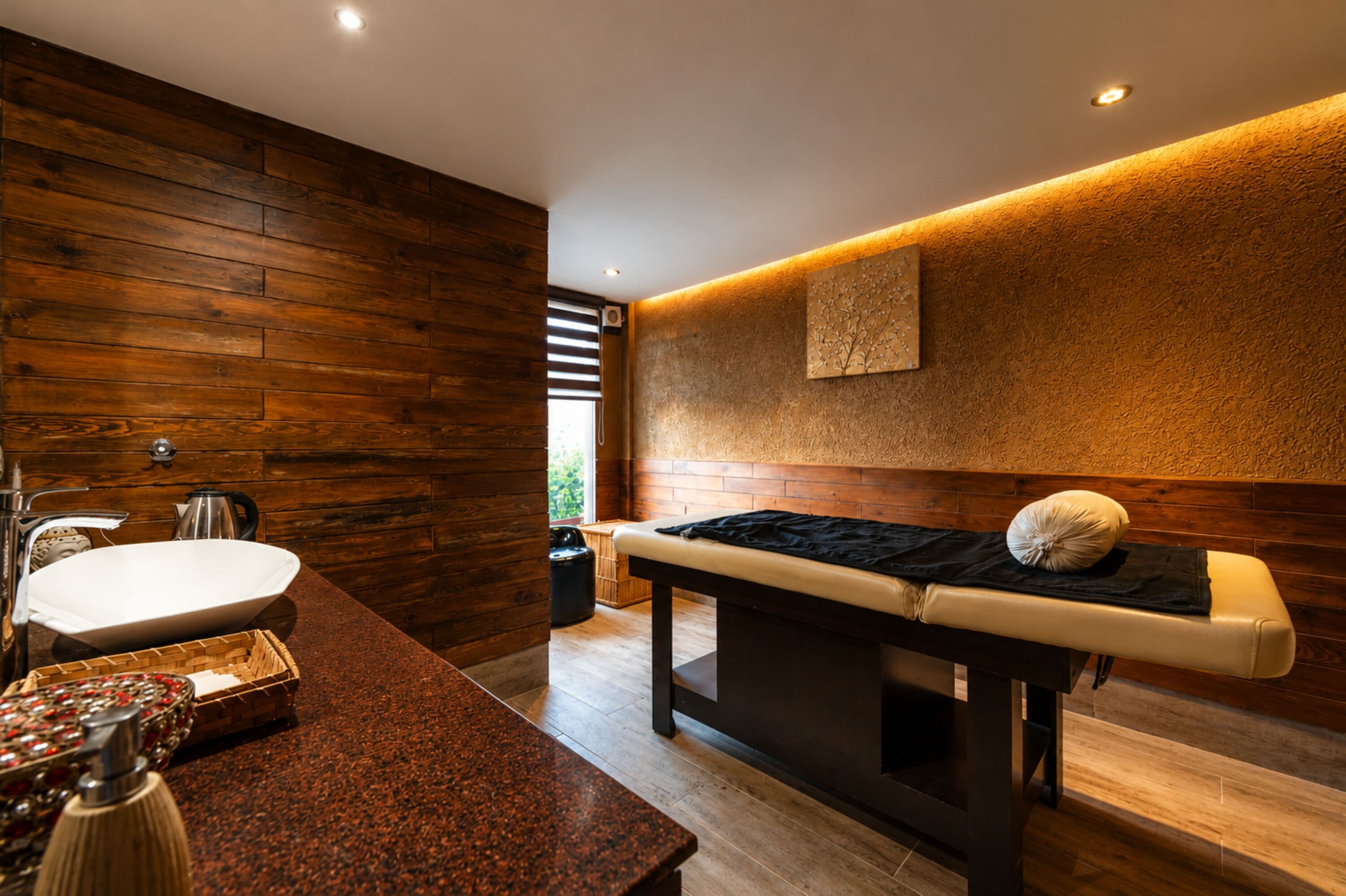 A modern spa room with a wooden wall, a massage table, a sink, and a decorative bowl.