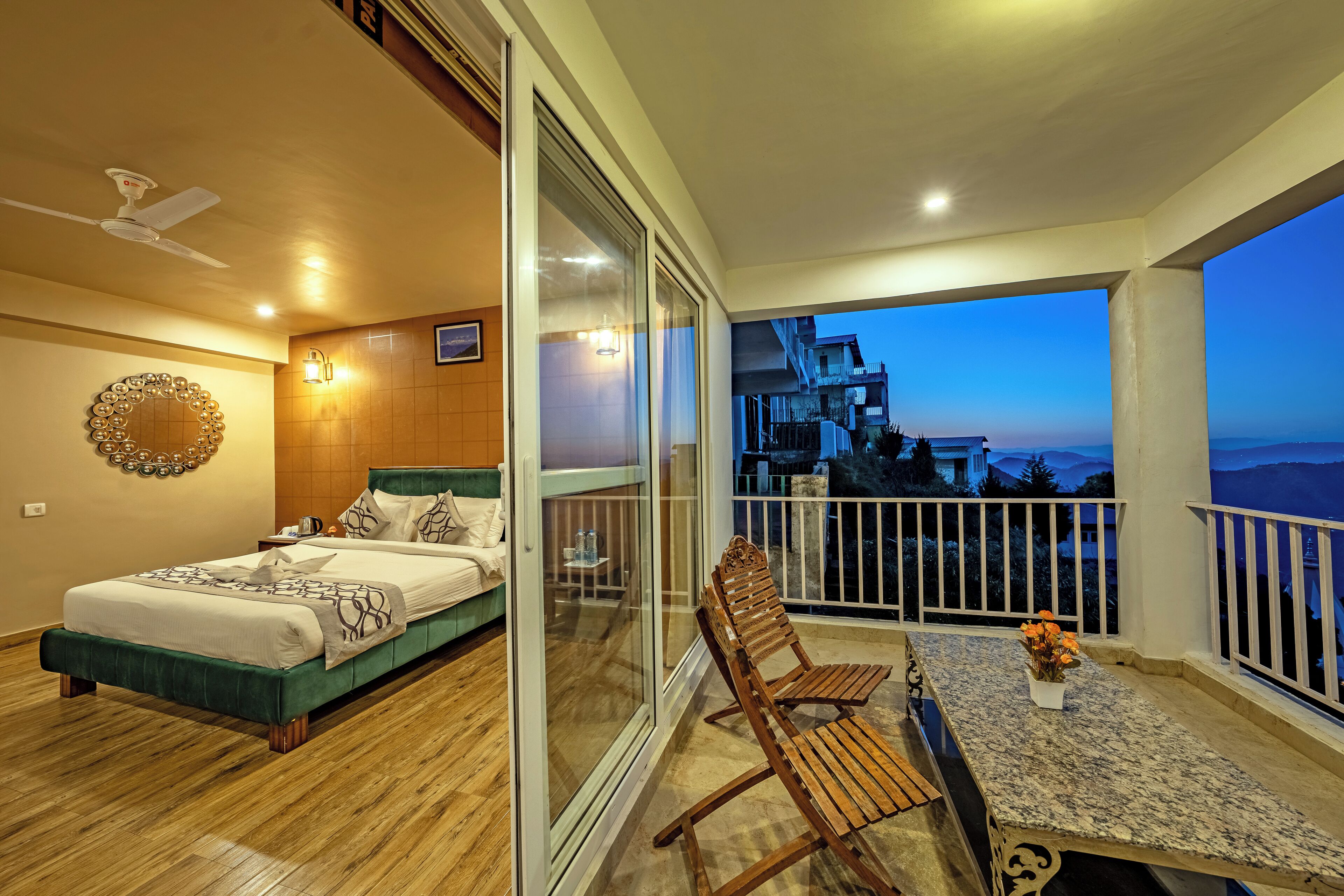A bedroom with a bed, a nightstand, and a bedside table. There is a balcony with a table and chairs, and a view of the cityscape.