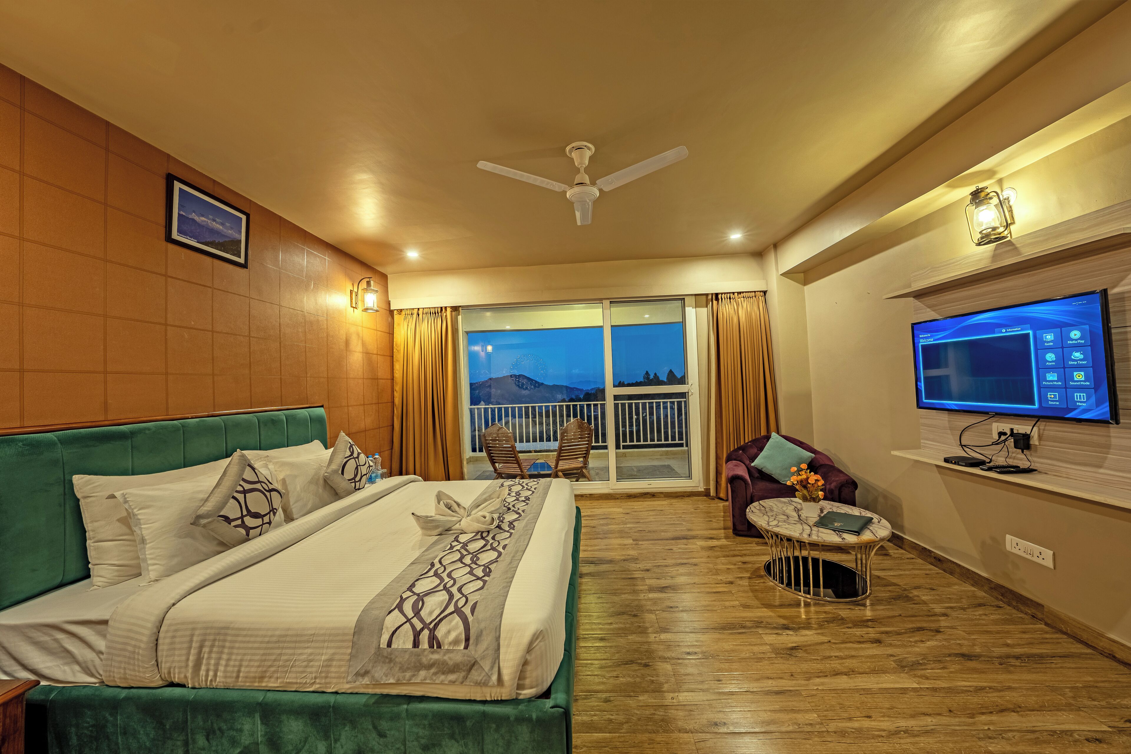 A hotel room with a large bed, a television, a balcony with a view, and a ceiling fan.