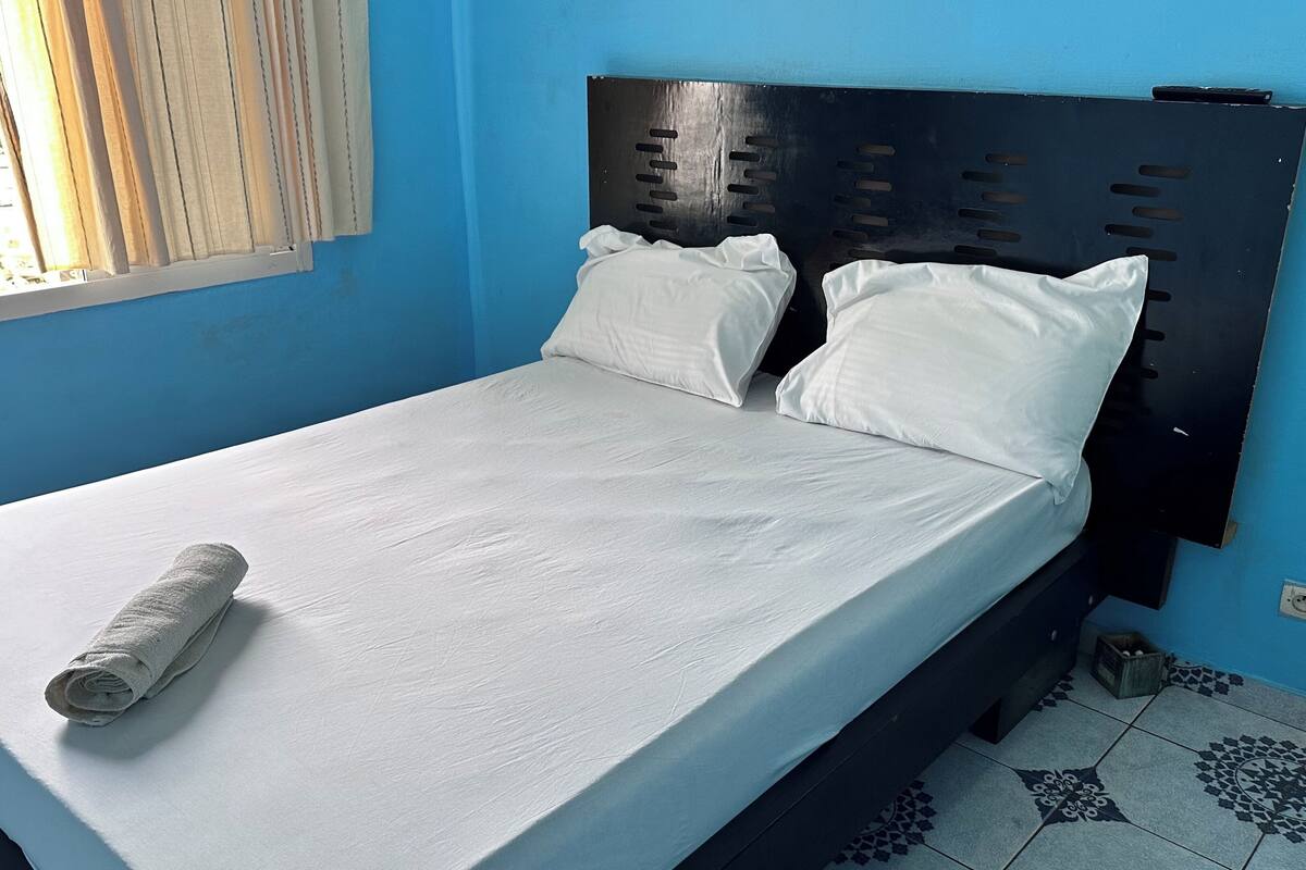 Un lit simple avec un matelas et des oreillers blancs, une serviette blanche enroulée et une tête de lit noire.