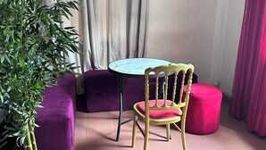 Een kleine kamer met een ronde tafel, een paarse bank, een rode poef en een kamerplant.