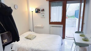 Een slaapkamer met een bed, een nachtkastje met een kop en schotel, een radiator, een raam met gordijnen, een wandklok en een jas aan de deur.