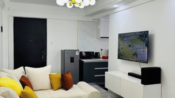 A modern living room with a sofa, a television, and a kitchen area in the background. - Cosy apartment in KILIMANI 
Mini golf 
restaurant 
Smrt TV
parking 
gym
security (Nairobi)